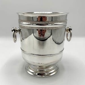 May include: A silver wine bucket with a polished finish. The bucket has a wide rim, a beaded band, and two ring handles. It sits on a pedestal base, reflecting light. A classic piece for serving chilled beverages.