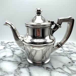 May include: A silver teapot with a rounded body and a lid with a knob on top. The teapot has a handle on the side and a spout on the front. The teapot has a circular embossed design on the front.