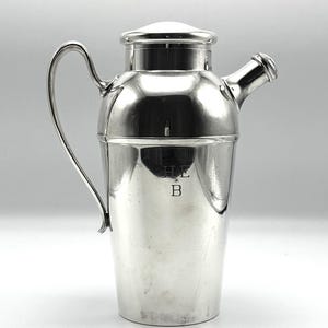 May include: A polished silver-toned cocktail shaker with a curved handle, a tapered body, and a removable cap. The front of the shaker features an engraved monogram with the letters H and E positioned above the letter B, separated by a small star.