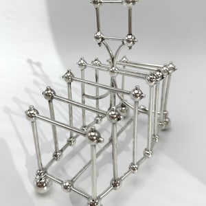 May include: A silver-toned metal toast rack featuring a series of vertical dividers connected by horizontal bars. The structure is composed of thin rods with small spherical finials at each joint and a central arched handle for carrying.