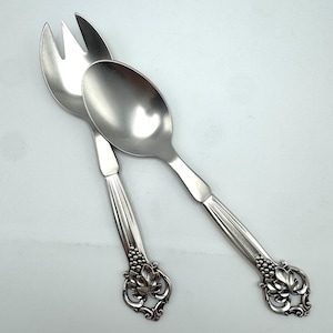 May include: A silver-toned metal salad serving set consisting of a three-pronged fork and a spoon. The handles feature a vertical ribbed design and a decorative base with embossed grape clusters and leaf motifs in a polished finish.