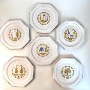 May include: A set of six white octagonal ceramic plates, each featuring a central circular illustration in shades of mustard yellow, brown, and blue. The designs depict various landscapes including a tower, a church, a house, trees, and a bridge.