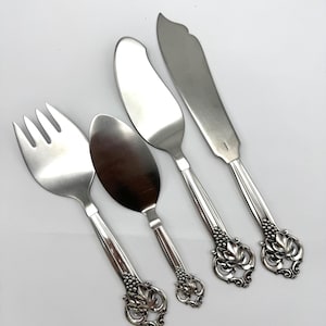 May include: A set of four silver-toned serving utensils including a wide fork, a rounded spoon, a curved spreader, and a flat-edged knife. Each handle features a detailed embossed design of grape clusters and leaves at the base.