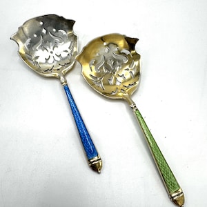 May include: Two ornate silver serving spoons with pierced floral designs. One spoon has a blue handle, the other a green handle. Both handles have gold accents and a pointed end. The spoons are likely for serving tea or sugar.