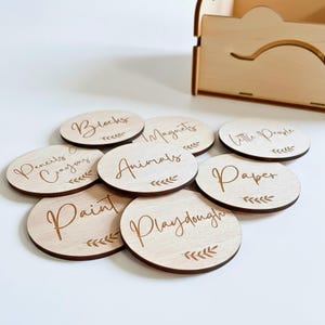 Puede incluir: Un conjunto de etiquetas ovaladas de madera con palabras grabadas con láser para organizar juguetes y materiales de manualidades. Las etiquetas incluyen "Blocks", "Magnets", "Little People", "Pencils & Crayons", "Animals", "Paper", "Paint" y "Playdough". Cada etiqueta tiene un diseño de hojas.