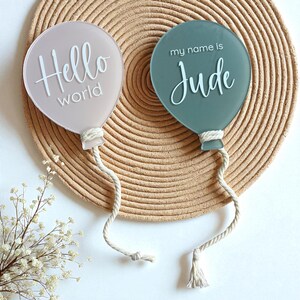 May include: Two balloon-shaped acrylic signs. One is light pink with the words "Hello world" in white script. The other is teal with "my name is Jude" in white script. Both have a white rope and tassel attached.
