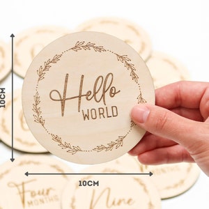 May include: A set of 10 cm round wooden baby milestone discs with a floral wreath design. One disc says "Hello World" in a script font.