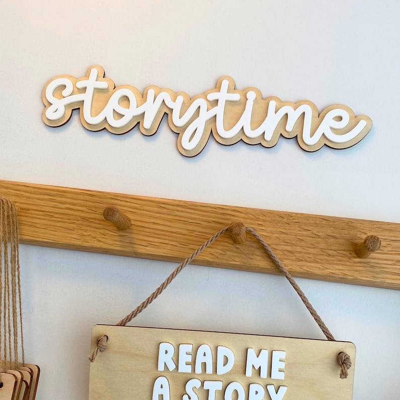 May include: A wooden sign that says "storytime" in a cursive font, mounted above a wooden peg rack. Below, a wooden sign with the text "READ ME A STORY" hangs from the rack. The signs are against a white wall.