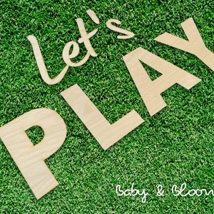 May include: Wooden letters spelling "Let's Play" on a green grass background. The letters are in a cursive font and are arranged in a diagonal line. The text "Baby & Bloom Co." is visible in the bottom right corner of the image.