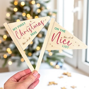 May include: Two wooden pennant flags. One reads "MY FIRST Christmas 2025" in green and red script, with a sleigh illustration. The other says "ON THE Nice LIST" in red script and green text, with a sleigh illustration. The flags are held in front of a blurred Christmas tree.