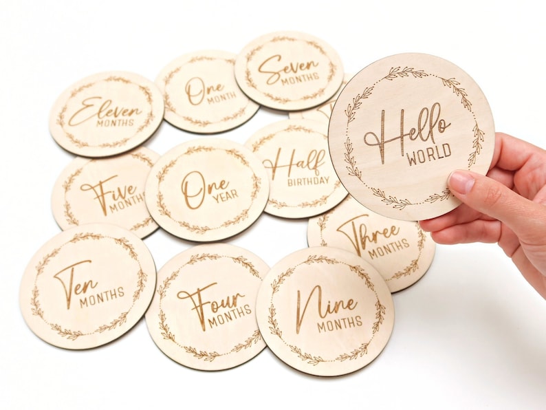 May include: A set of 11 wooden baby milestone discs with gold lettering. The discs feature phrases like "One Month", "Three Months", "Half Birthday", "One Year", and "Hello World".