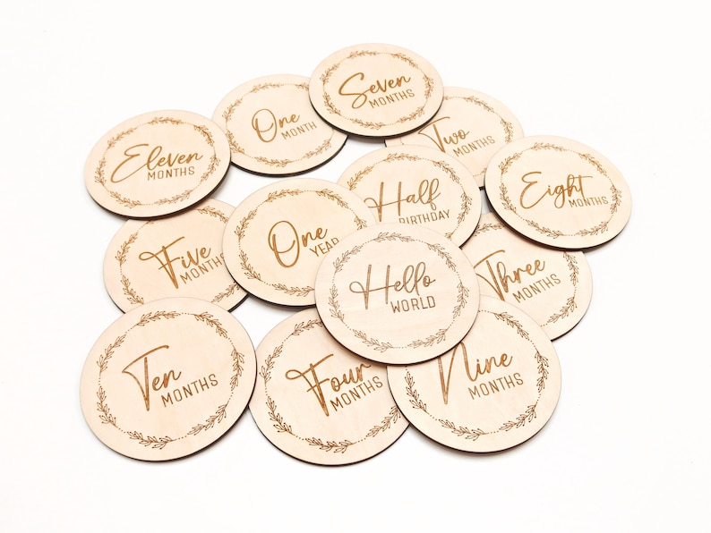 May include: A set of 12 wooden baby milestone discs with gold lettering. The discs feature phrases like "One Month", "Five Months", "Half Birthday", "One Year", and "Hello World".