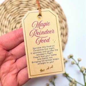 Puede incluir: Una etiqueta de madera con las palabras "Magic Reindeer Food" en escritura roja. La etiqueta incluye texto sobre cómo espolvorear la comida en Nochebuena. Una pequeña ilustración del trineo y los renos de Papá Noel está en la parte inferior.