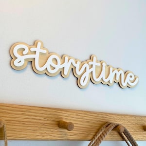 Puede incluir: Un letrero de madera en capas con la palabra "Storytime" en letras blancas sobre madera clara. El letrero está fijado a una pared blanca encima de un perchero de madera.