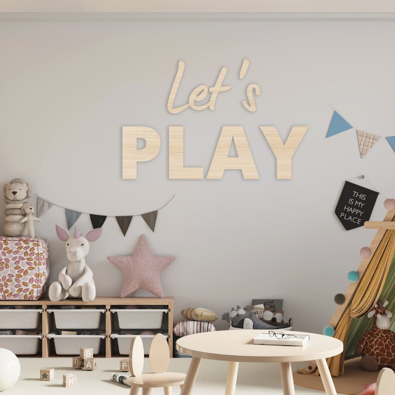 May include: A light wood playroom sign with the words "Let's Play" in a whimsical font. The sign is hanging on a white wall with a colorful garland of flags hanging below it. There is a small wooden table and chairs in the foreground, and a teepee tent in the background.
