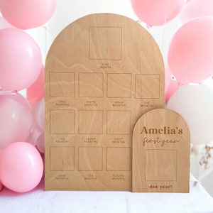 May include: Wooden milestone sign with 12 squares for each month of a baby's first year. The sign is decorated with the text "Amelia's first year" and "one year!" in a brown font.