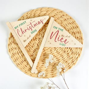 May include: Two wooden pennant flags with text for Christmas. One flag reads "MY FIRST Christmas 2025" in red script, with a sleigh and reindeer graphic. The other flag says "ON THE Nice LIST" in red script, with a sleigh and reindeer graphic.