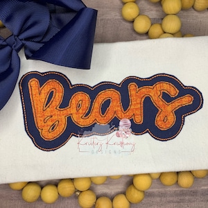 Pode incluir: Um tecido branco com um laço azul marinho no topo. O tecido tem um contorno azul marinho com um texto laranja "Bears" no interior. O texto tem um aspeto texturizado. O tecido está sobre uma superfície de madeira com uma corda de contas amarelas.