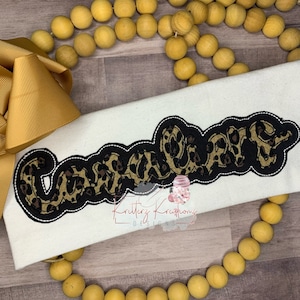 May include: A white fabric with the word "Caramel" written in black outlined letters with a leopard print pattern inside the letters.