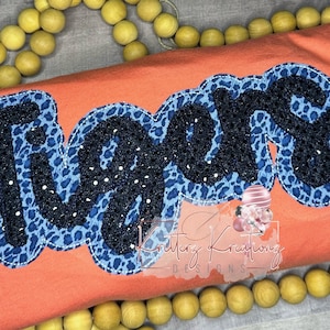 以下が含まれることがあります： コーラルオレンジのTシャツに、青と黒の「タイガース」のデザインが施されています。デザインは白で縁取られており、レオパード柄になっています。