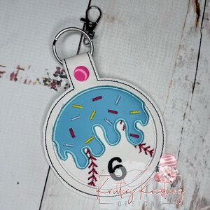 May include: White key chain with a blue and white donut design. The donut has sprinkles and a pink center. The number 6 is in the center of the donut.