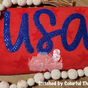 Könnte beinhalten: Rotes Shirt mit einem blauen, mit Pailletten besetzten "USA"-Design. Das Shirt hat ein Muster aus weißen Sternen.