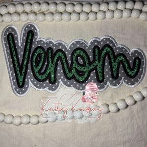 May include: A fabric applique with the word "Venom" in green glitter lettering, outlined in black and gray, with a white polka dot background. The applique is on a cream-colored fabric, with white beads at the top and bottom.