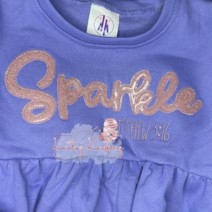 Puede incluir: Sudadera morada con un diseño de purpurina rosa que dice "Sparkle" y "Mateo 5:16".