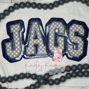 May include: A gray and blue embroidered patch with the word "JAGS" in a varsity style font. The letters are outlined in blue and have a white polka dot pattern.