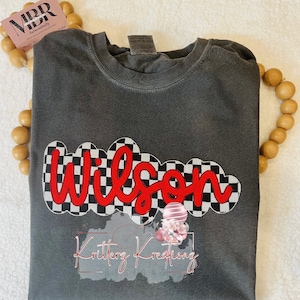 May include: A dark gray t-shirt with a red and black checkered design featuring the name "Wilson" in red lettering. The shirt also has a small pink and white balloon graphic with the text "Kritterz Kreations" underneath.