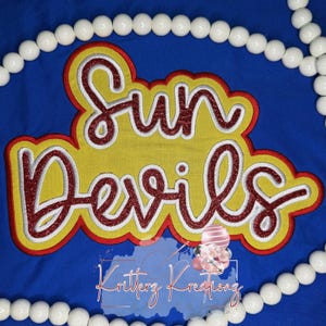 以下が含まれることがあります： 赤のグリッター調の筆記体でSun Devilsと刺繍されたワッペン。白と黄色の縁取りが施され、無地の青い布地の上に配置されています。ワッペンの周囲は、円形に並べられた白いビーズの列で囲まれています。