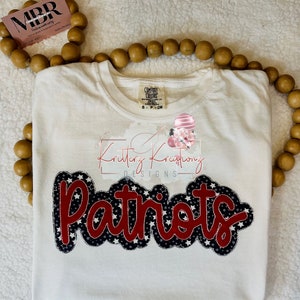 Peut inclure: T-shirt blanc avec un motif "Patriots" rouge et bleu avec des étoiles. Le motif est bordé de noir et a un aspect cousu. Le t-shirt porte une étiquette Comfort Colors et les mots "Kritterz Kreations Designs" sont imprimés sur le t-shirt.