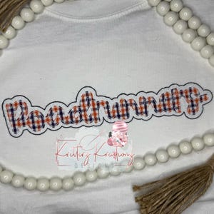 May include: White t-shirt with the embroidered word "Roadrunner" in a plaid pattern of red, white, and blue. The shirt also features the logo "Kritter Kreations Designs" and is decorated with white beads and a brown tassel.