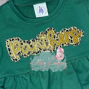 Könnte beinhalten: Grünes Langarmshirt mit einem gold-schwarzen "Panthers"-Design mit Leopardenmuster. Das Shirt hat einen gerafften Saum.