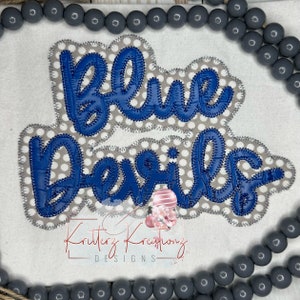 Może przedstawiać: Aplikacja "Blue Devils" na szarym i białym tle w kropki. Aplikacja jest wykonana z niebieskiego materiału o fakturowanej powierzchni. Tekst jest napisany kursywą.