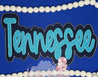 May include: A blue fabric patch featuring the word Tennessee in bright turquoise script with white stitching. The text is layered over a dark navy blue scalloped background. The patch is positioned between two rows of white wooden beads.