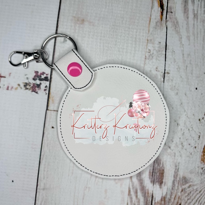 May include: White round keychain with a pink button and a pink and white design that says "Kritterz Kreations Designs".
