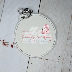 May include: White round leather keychain with a black stitched border. The keychain features a pink and white balloon design with the text "Kritterz Kreations DESIGNS".
