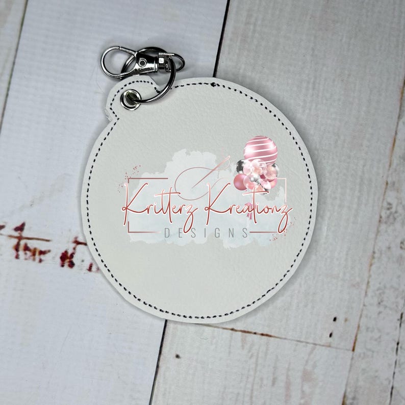 May include: White round leather keychain with a black stitched border. The keychain features a pink and white balloon design with the text "Kritterz Kreations DESIGNS".