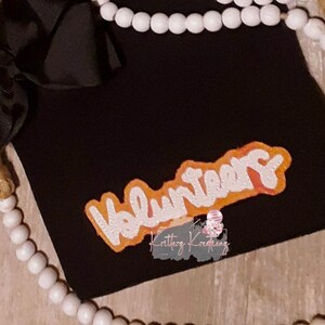 May include: A black t-shirt with an orange and white "Volunteers" graphic. The graphic is outlined in orange and has a white fill.
