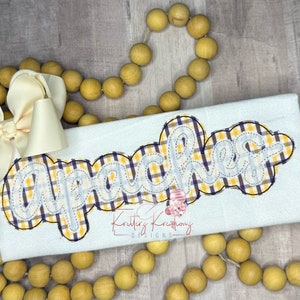 May include: A white t-shirt with the word "Apaches" embroidered in yellow and purple plaid fabric. The word is outlined in black and has a white outline.