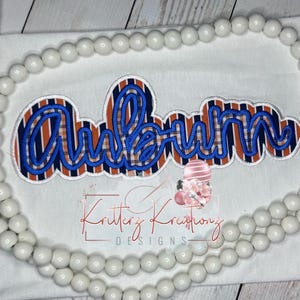 May include: White fabric with the word "Auburn" embroidered in blue script over a plaid pattern of orange, navy, and white. The text "Kritterz Kreazionz Designs" is printed below the word "Auburn". A string of white beads frames the top and sides.