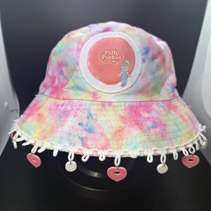 May include: A tie-dye bucket hat with a Polly Pocket logo patch. The hat features a white lace trim with pink heart and white circle charms. The hat's colors include pink, yellow, blue, and green.