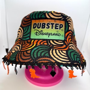May include: A black bucket hat with a colorful pattern of orange, green, and brown swirls. The hat has a black label with the text "Dubstep Disneyland" on it. The hat has a black trim with orange and black dangling charms.