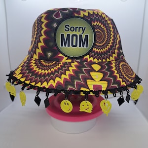 May include: A yellow, red, and black bucket hat with a black trim and a yellow smiley face charm. The hat has a round patch on the front that says "Sorry MOM".