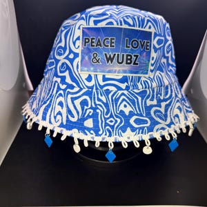 May include: A blue and white patterned bucket hat with the text "PEACE LOVE & WUBZ" on the front. The hat has a white trim with blue diamond-shaped charms hanging from it. The hat is a stylish accessory.