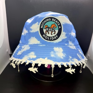 May include: Blue bucket hat with a cloud pattern and a patch that reads "NOTHING BEATS A HOLIDAY". The hat has a white lace trim with airplane and spoon charms. A stylish accessory for travel.