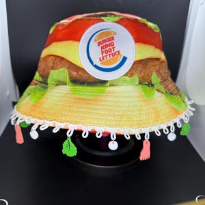 Puede incluir: Un sombrero de cubo amarillo, rojo y verde con un logotipo de Burger King Foot Lettuce. El sombrero tiene un borde de encaje blanco con abalorios rosas, verdes y blancos.