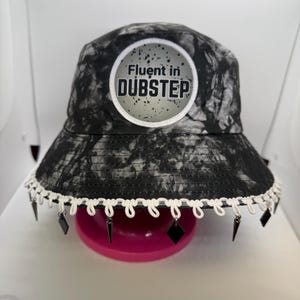 Puede incluir: Sombrero de cubo de teñido anudado en blanco y negro con un parche circular blanco que dice "Fluent in Dubstep". El sombrero tiene un borde blanco y abalorios negros colgantes.
