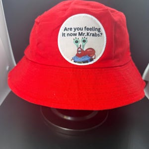 Puede incluir: Sombrero de pescador rojo brillante con un parche circular con Mr. Krabs de Bob Esponja. El parche tiene el texto "Are you feeling it now Mr. Krabs?" en fuente negra. El sombrero est&aacute; hecho de tela suave.